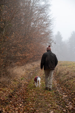 Mann mit Pudelm&uuml;tze und Hund mit Strickpulli im Partnerlook stapfen in den nebelverhangenen Herbstwald