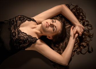 Artistic portrait of young woman in black lace robe lying on floor with arms raised above head. Her...