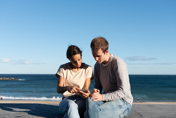 Young couple sitting on a stone seat and checking their smartphone at the seaside in a sunny day
