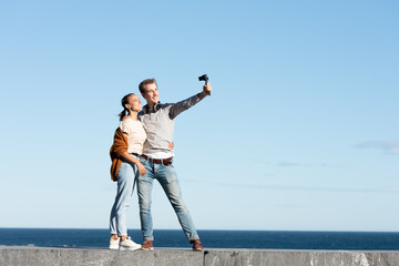 Social media content creation concept. A young couple taking themselves a selfie with a compact camera in a sunny day at the seaside.