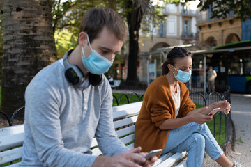 Obraz premium Young man and woman sitting on a bench checking their phones while keeping social distance during Coronavirus pandemic. Couple wearing face masks
