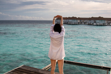 Beautiful traveler woman in a beach dress, standing with morning sun. Young woman in white clothes stretching after getting up from bed. Romantic honeymoon getaway in overwater bungalows villa.