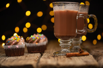 Glasses with hot latte and chocolate cupcakes on rustic wooden table served with cinnamon stick on black background with yellow lights. Luxury dessert in cafe. Delicious food. Bakery concept.