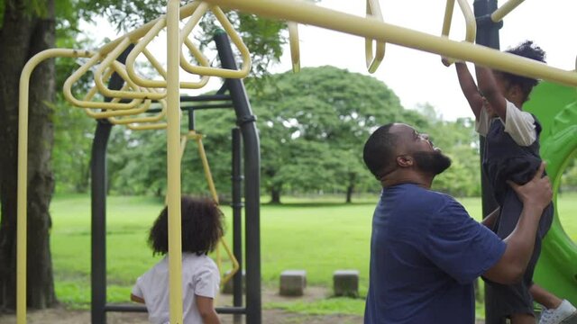 Happy affectionate mixed race family. African man father carrying little daughter playing at playground in the park. Dad and two child girl enjoy and having fun together in outdoor weekend vacation.