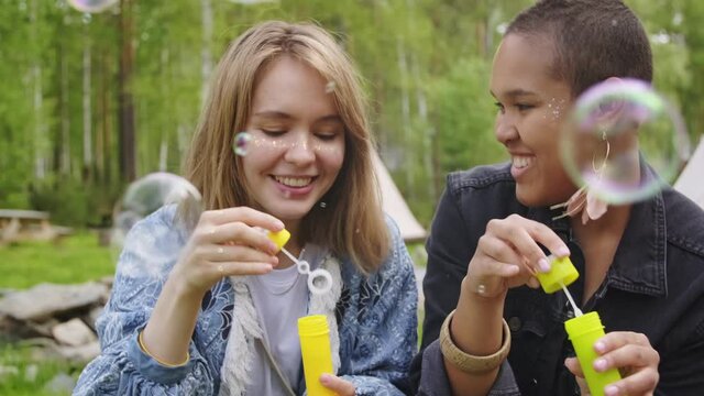 Closeup of young cheerful girls blowing soap bubbles together laughing and catching positive vibes outdoors at campsite