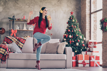 Photo of pretty young lady dance hold smartphone sing raise knee wear earphones red sweater in decorated living room indoors