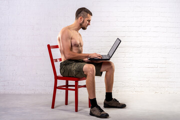 A man with a naked torso sits on a chair and holds a laptop.