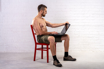 A man with a naked torso sits on a chair and holds a laptop.