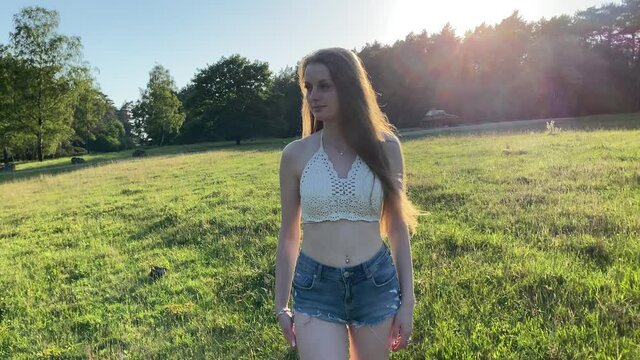 H&uuml;bsche junge Frau spaziert &uuml;ber eine Wiese im Park an einem warmen sonnigen Abend im Sommer