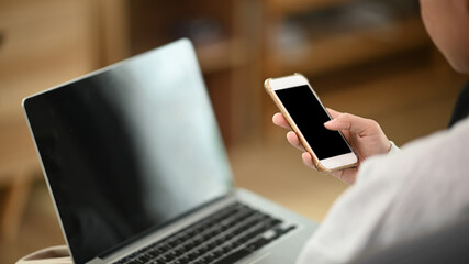 Cropped shot of female using mock up smartphone while working with mock up laptop in office.