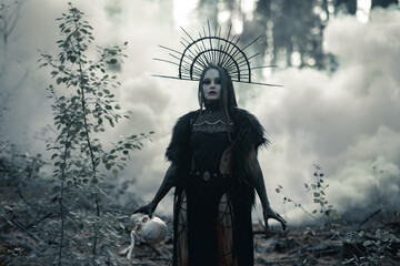 Woman in image of witch walks with human skull in through smoke in forest.