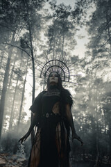 Woman in image of witch stands in penetrated by the sun rays foggy forest.