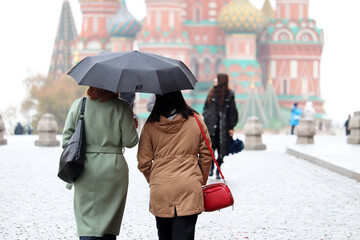 Rain in a Moscow, two women in autumn coats with one umbrella walking on the Red Square on background of St. Basil's Cathedral. People in rainy weather, fall season