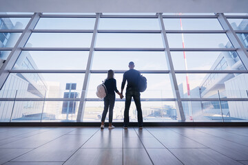 Traveling concept. Back view of loving couple in casual wear standing near the window of international airport terminal.