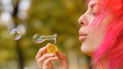 Profile of happy adult girl blowing soap bubbles front camera on nature. Close-up. Good-looking girl blows bubbles at camera and has fun. Portrait young cheerful caucasian woman with bright pink hair.