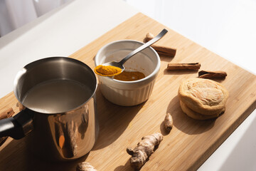 Cutting board with curcuma powder in white bowl,  pot with almond milk, cinnamon sticks and row turmeric roots on wooden background. Preparation steps of homemade curcuma latte.