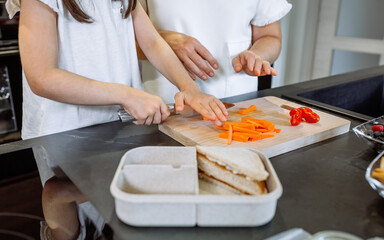 Unrecognizable mother teaching her daughter to cut vegetables to prepare the lunch box for school