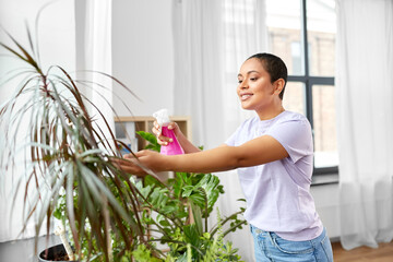 people, housekeeping and plants care concept - african american woman spraying houseplant with...