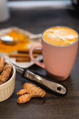 Curcuma latte in pink mug with curcuma roots, cinnamon sticks at table. Homemade Golden Latte