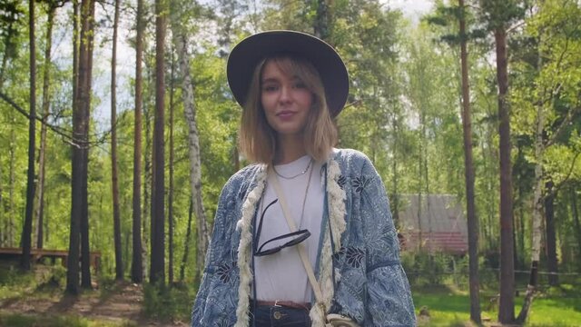 Tracking medium footage of attractive stylish caucasian young woman dressed in boho style clothes looking at camera standing among trees in forest in daylight
