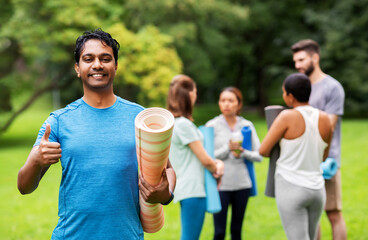 Fototapeta na wymiar fitness, sport and healthy lifestyle concept - happy smiling indian young man with mat showing thumbs up over group of people meeting for yoga class at summer park