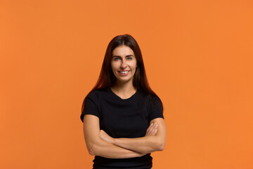 Obraz premium this is a success. Studio shot of confident brunette caucasian woman in black t-shirt keeps hands crossed, has long hair, looks directly at camera with gentle smile. Isolated over an orange wall