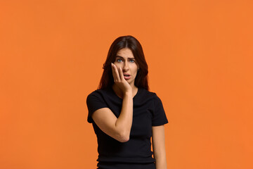 I hate to even watch. Shocked caucasian woman in black t-shirt disgusting expression as sees something unpleasant, keeping palm on her cheek. Female isolated over an orange wall