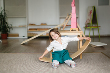 child girl in a linen apron sways on balancing board, developing sensory and sports activities in montessori and earlier child development, children's independence - obrazy, fototapety, plakaty