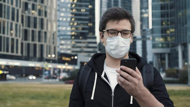 Man wearing a mask walking with a phone on background of skyscrapers in business centre. Man dressed in casual clothes holding a phone while walking 