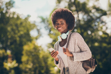 Photo of positive dark skin lady use smartphone in autumn outside park walk wear cardigan rucksack