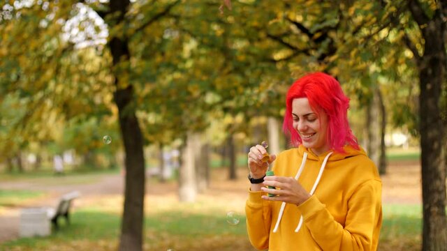 Happy adult girl blowing soap bubbles  on nature. Good-looking girl blows bubbles at camera and has fun. Portrait young cheerful caucasian woman with bright pink hair. 