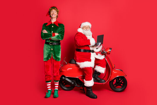 Full length photo of two men elf folded arms santa sit moped hold netbook wear x-mas costume coat cap boots isolated red color background