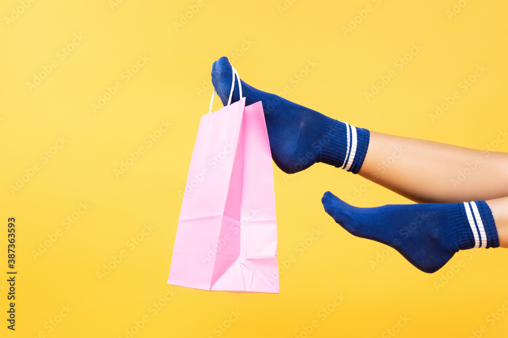 Poster Cropped view of pink paper bag hanging on woman leg in socks isolated on yellow background