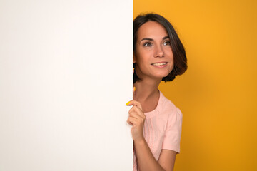 Young woman looks up holding white empty blank with copy space on it. Isolated on yellow background. Studio shot.