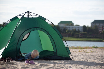 Small child lies in tourist tent against river and houses background. Camping with children. Family vacation