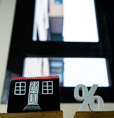View from below of a miniature house and a percentage symbol on some woods with a window in the background. It can represent any real estate or property concept or even family or industry crisis