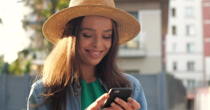 Beautiful Latino American Female is browsing her Phone outdoor, smiling Charmingly. Stylish Girl in Hat is texting, browsing, using her Smartphone on a warm sunny Day. Positive Emotions. Apps.