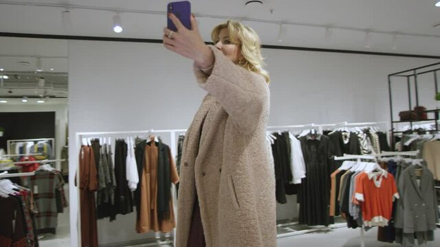 Aged woman is taking a photo of herself dressed in a fur coat at clothing store, tracking shot, 360 degree