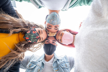 Group of four teenagers looking down arm in arm wearing protective mask - Friends looking at camera and laughing hugging each other in covid-19 pandemic period