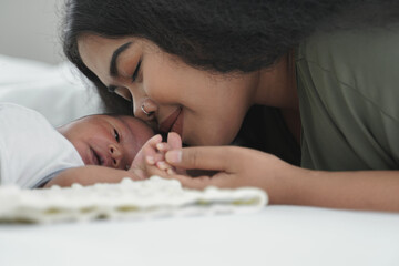 African mother kissing her new born baby on a white bed and holding little infant boy's hand while he is sleeping on bed at bedroom