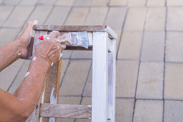 Senior man holding brush for paint white on old wooden chair without a backrest Hand of man. Old chairs are painted white. Renovation of old furniture