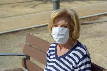 Elderly junior woman holding a sanity mask with blonde hair and blue eyes sitting on a bench. 