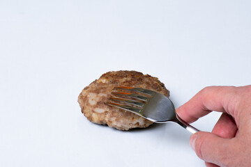 Hand holds a fork over cooked meat patty on a white plate isolated on white background, close up, copy space