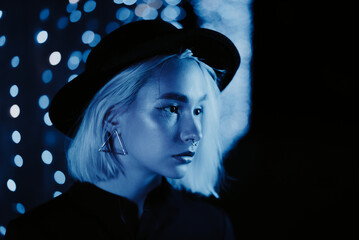 Millennial enigmatic pretty girl blond hairstyle near blue glowing neon wall at night. Hair, hipster hat, nose piercing. Mysterious woman