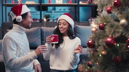 African american couple holding sparklers and cup near christmas tree - Powered by Adobe