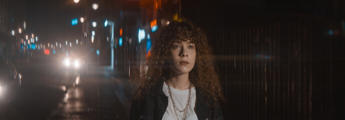 Curly haired woman in white t-shirt and black jacket walks street at the night in anamorphic flares and city lights - Fashion clothing and feminism concept