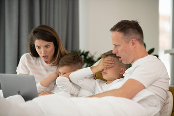 Fototapeta premium Beautiful parents with kids enjoying at home. Young family watching movie on lap top.