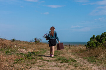 Naklejka na ściany i meble Young woman with long brown hair in denim jacket, black skirt, vintage suitcase,purple wild flowers bouquet off-road sea landscape. Hitchhiker on countryside trip. Lifestyle photo lady walking outdoor
