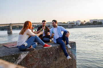 Group of young people sitting by the river .They joying in city view . Young man playing guitar while his friends drinks beer .	

