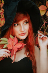 Halloween Beautiful young Witch girl in witches hat on spooky dark magic forest background. Portrait of model girl with autumn bright leaves, Fall look style. redhead girl with big eyes magical sight